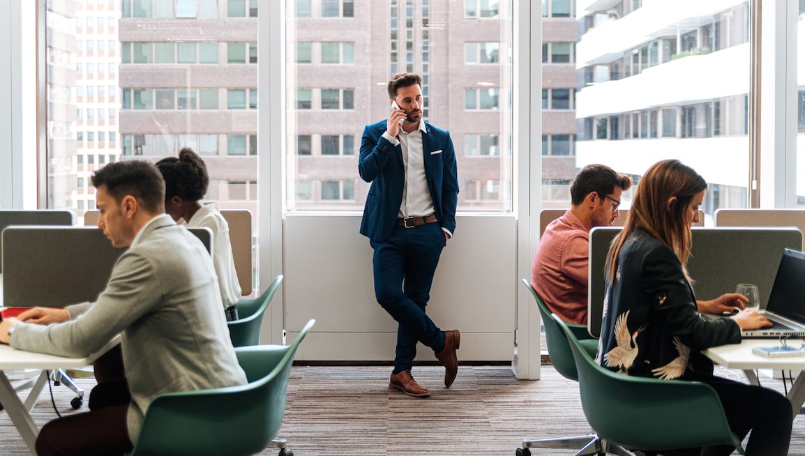 man-in-suit-stands-between-tables-of-people-working